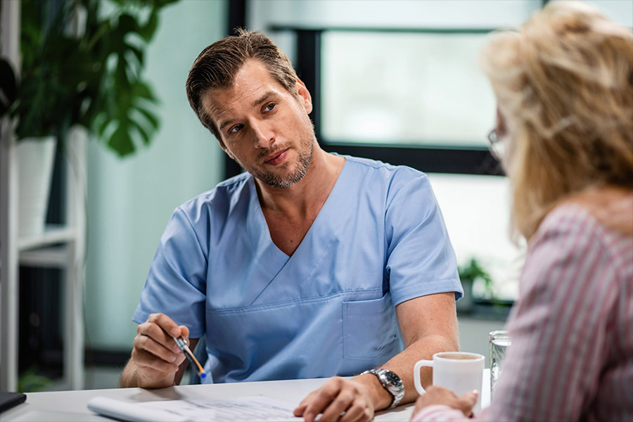 Doctor in scrubs talking to a patient across a desk during a consultation.